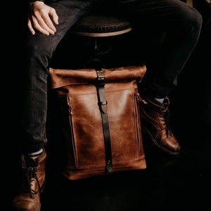 May include: A brown leather backpack with a roll-top closure and black leather straps. The backpack is sitting on a stool.