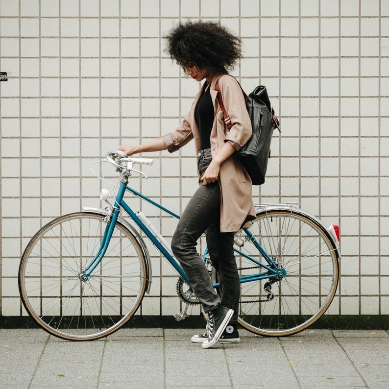 May include: A woman with curly brown hair is standing next to a blue bicycle with a black leather rucksack. She is wearing a beige jacket, black trousers, and black trainers.