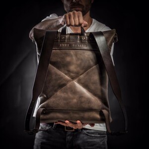 May include: A brown leather backpack with a flap top and adjustable straps. The backpack has a stitched diamond pattern and the text "KRUK GARAGE" embossed on the front.