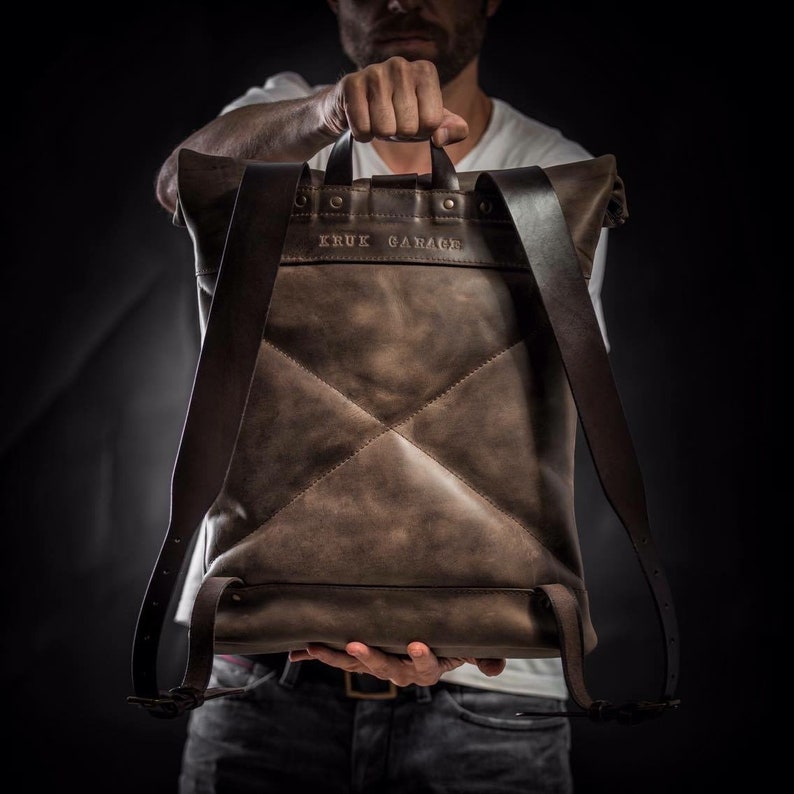 May include: A brown leather backpack with a flap top and adjustable straps. The backpack has a stitched diamond pattern and the text "KRUK GARAGE" embossed on the front.