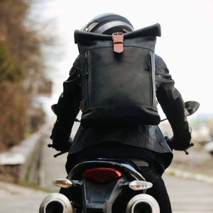 May include: A person wearing a black leather jacket and a black leather backpack with a brown leather strap rides a motorcycle. The backpack has a roll-top closure.