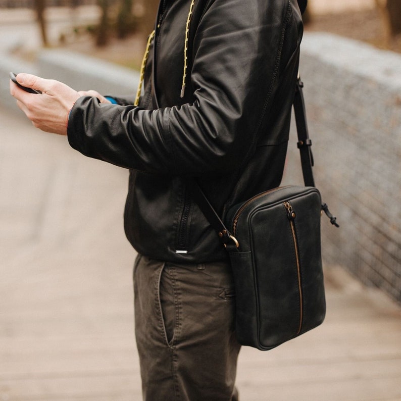 May include: A black leather crossbody bag with a zip closure and a long adjustable strap. The bag is being worn by a person wearing a black leather jacket and brown trousers.