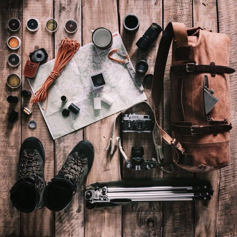 May include: An overhead view of travel and photography equipment on a wooden surface. The image features a brown leather backpack, hiking boots, cameras, a map, a tripod, and other accessories, arranged to suggest a journey.
