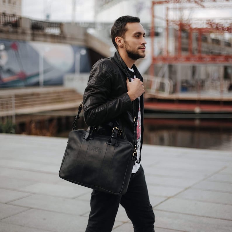 May include: A man wearing a black leather jacket and black jeans is carrying a black leather briefcase with a gold buckle. The briefcase has the text "FREEDOM" embossed on the front.