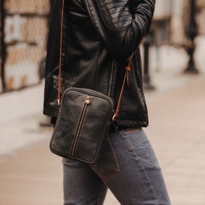 May include: A black leather crossbody bag with a brown strap. The bag has a zip closure and a flap with a buckle.