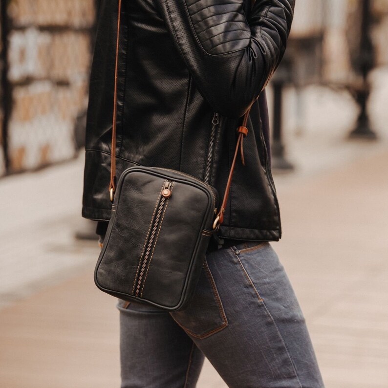 May include: A black leather crossbody bag with a brown strap. The bag has a zip closure and a flap with a buckle.