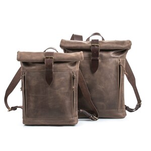 May include: Two brown leather roll-top backpacks with adjustable straps and buckles. The smaller backpack is in front of the larger backpack.