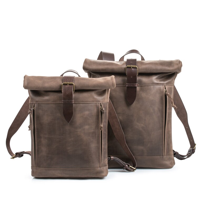 May include: Two brown leather roll-top backpacks with adjustable straps and buckles. The smaller backpack is in front of the larger backpack.