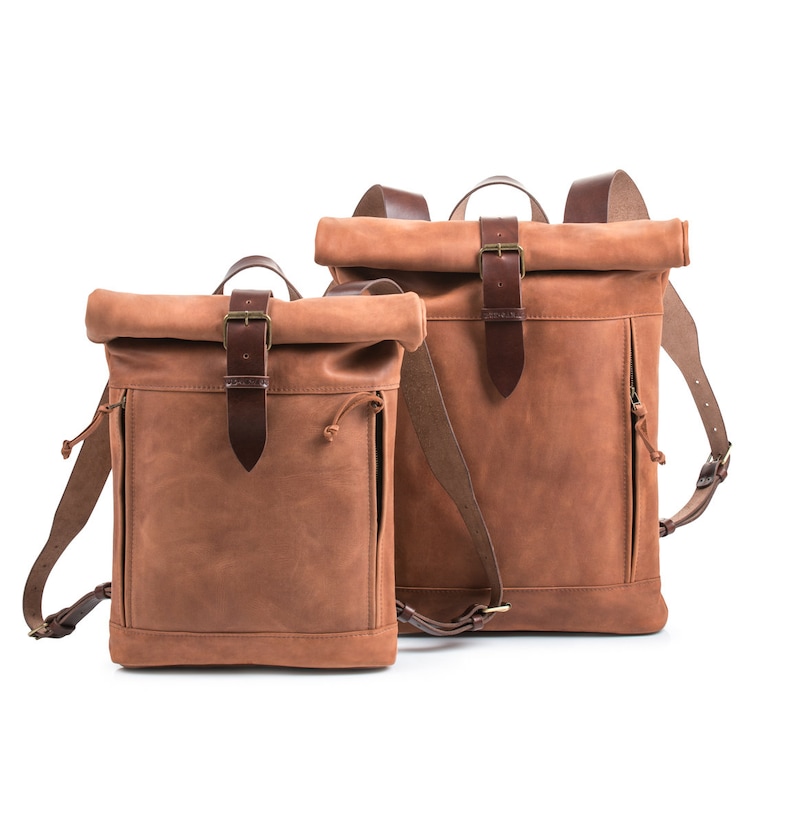 May include: Two brown leather roll-top backpacks with adjustable straps and buckles. The smaller backpack is in front of the larger backpack.