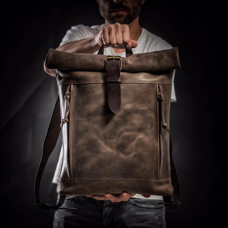 May include: A brown leather backpack with a roll-top closure and a buckle. The backpack has a distressed look and is made of high-quality leather.