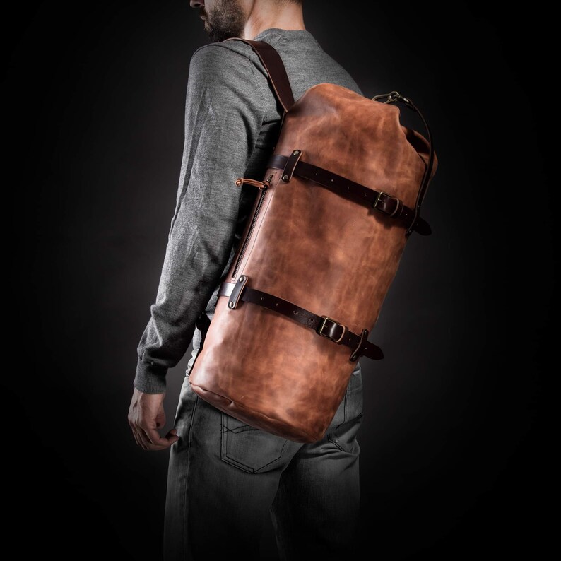 May include: A brown leather backpack with dark brown straps and buckles. The rectangular bag is worn over the shoulder. The leather has a distressed appearance, and the bag appears to be well-made. The bag is approximately 76 cm tall.
