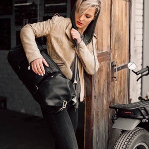 May include: A black leather duffel bag with silver fittings is held by a person. The bag has a cylindrical shape and a top handle. The person is wearing a tan quilted jacket and black jeans. A motorcycle is partially visible in the background.