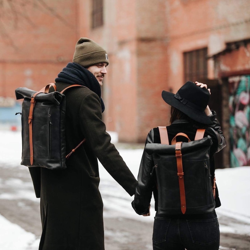 May include: Two people walking down a snowy street. The person in the front is wearing a black coat, a brown scarf, and a black leather backpack with brown straps. The person in the back is wearing a black leather jacket, a black hat, and a black leather backpack with brown straps.