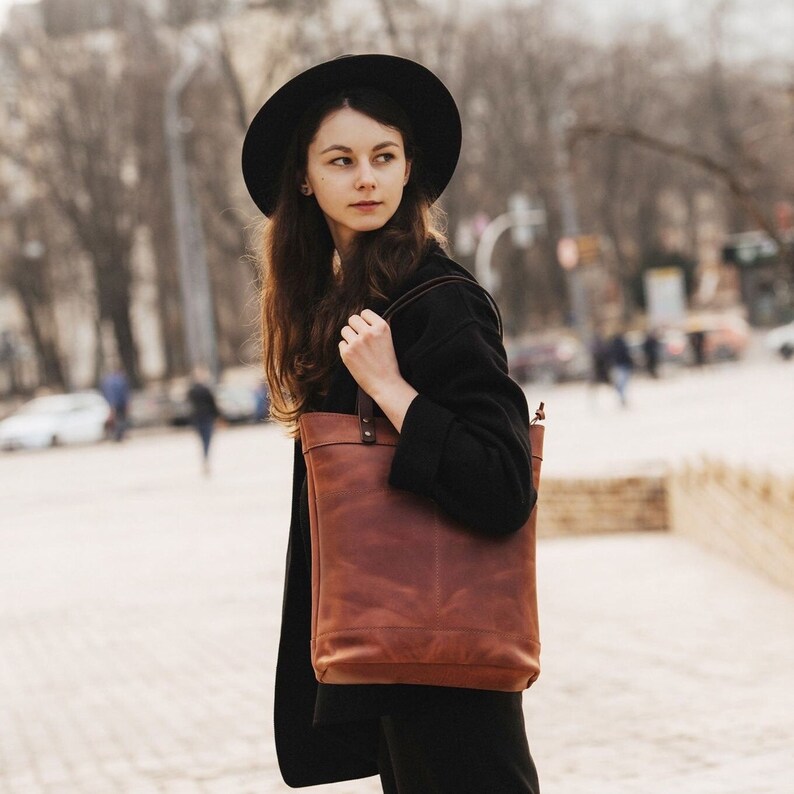 May include: A woman wearing a black hat and a black jacket is carrying a brown leather tote bag with a strap.