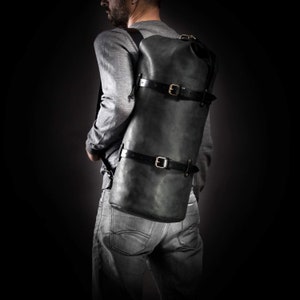 May include: A dark grey leather backpack with black straps and gold-coloured buckles. The cylindrical bag is worn over the shoulders, with two horizontal straps securing the main compartment. The background is a solid black.