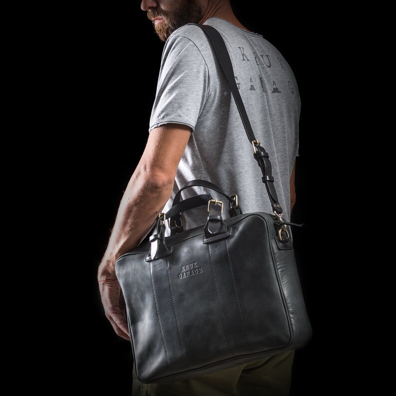 May include: A black leather satchel bag with a shoulder strap. The bag has a gold buckle and the text "KRUX GARAGE" embossed on the front.