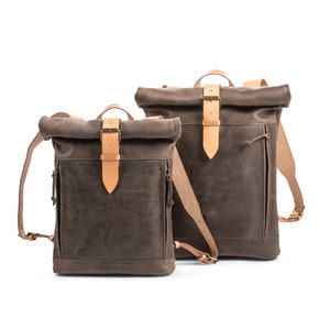May include: Two brown leather roll-top backpacks with tan leather straps and buckles. The larger backpack is behind the smaller backpack.