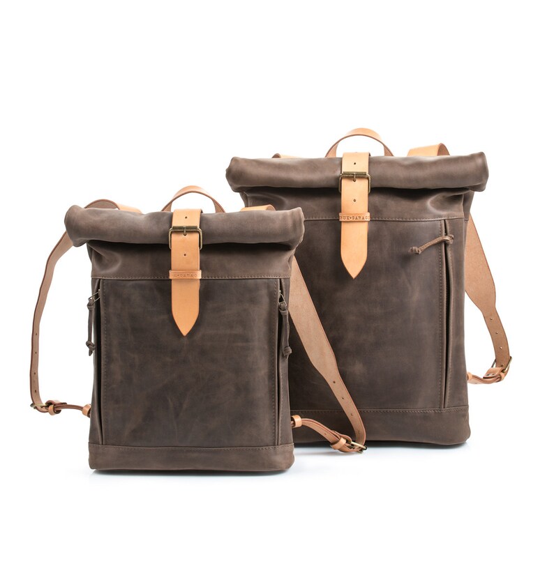 May include: Two brown leather roll-top backpacks with tan leather straps and buckles. The larger backpack is behind the smaller backpack.
