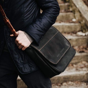 May include: A black leather messenger bag with a brown leather strap. The bag has a flap closure and a stitched design. The bag has the text "KNIX" printed on the flap.