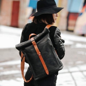 May include: A black leather backpack with a roll-top closure and brown leather straps. The backpack has a buckle on the front and a zipped pocket on the side.