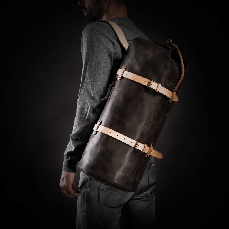 May include: A dark brown leather backpack with light tan straps and silver buckles. The cylindrical bag is worn over the shoulder, showcasing its design and craftsmanship. The bag is designed for travel and outdoor activities.
