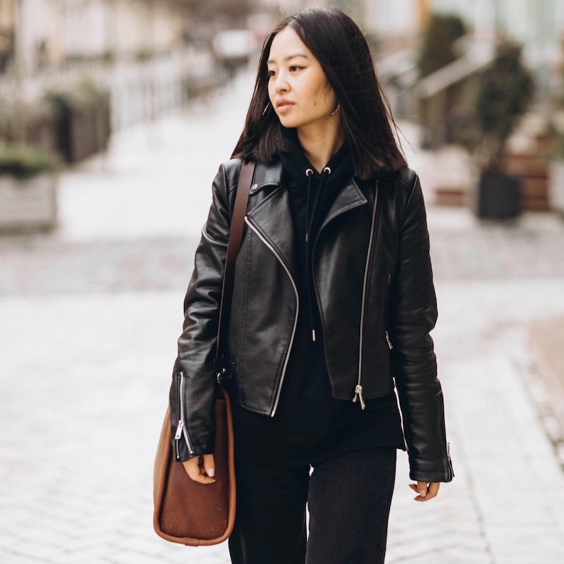 May include: A woman wearing a black leather jacket with a brown leather bag slung over her shoulder. She is walking down a street.