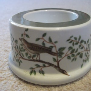 May include: White ceramic candle holder with a green and brown bird design. The holder has a wide, round opening at the top.