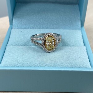 May include: A ring featuring a central oval-cut yellow stone set in yellow gold, surrounded by a halo of small white stones. The band is made of silver-toned metal and is split into two rows, also encrusted with small white stones.