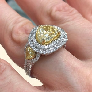May include: A pear-shaped yellow diamond ring with a double halo of smaller diamonds. The ring is set in white gold and features a smaller pear-shaped yellow diamond on the side.