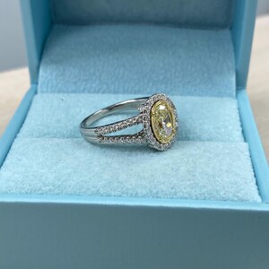 May include: A silver-toned ring featuring a central oval-cut yellow stone surrounded by a halo of small white stones. The band is split into two sections, both encrusted with small white stones, and is presented in a light blue box.