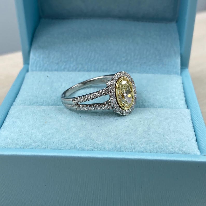May include: A silver-toned ring featuring a central oval-cut yellow stone surrounded by a halo of small white stones. The band is split into two sections, both encrusted with small white stones, and is presented in a light blue box.