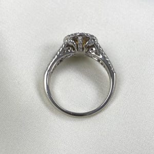 May include: A silver-toned ring featuring a central yellow stone surrounded by a halo of small clear stones. The band and setting are decorated with intricate cut-out metalwork and additional small clear stones along the shoulders.