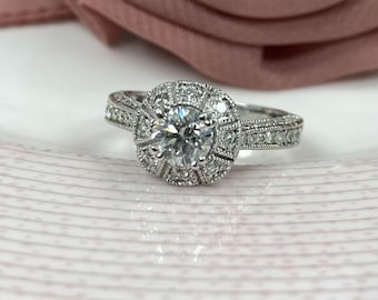 Könnte beinhalten: Ein silberner Verlobungsring mit einem großen runden Diamanten, der von kleineren Diamanten umgeben ist. Der Ring hat ein zartes Band mit einem Milgrain-Rand.