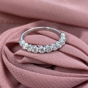 May include: A delicate silver band with a row of sparkling round diamonds set in a half-circle shape. The band is on a pink fabric background.