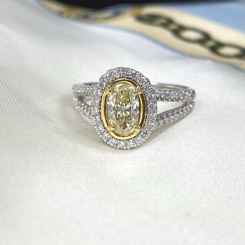 May include: A ring featuring a central oval-cut yellow diamond set in a yellow gold bezel, surrounded by a halo of small white diamonds. The split shank band is crafted from silver-toned metal and is encrusted with small white diamonds.