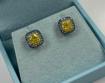 May include: A pair of yellow and blue diamond earrings in a blue velvet box. The earrings are square shaped with a yellow diamond in the center and a halo of blue diamonds around it.