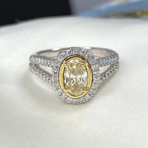 May include: A ring featuring a central oval-cut light yellow stone set in a yellow gold bezel, surrounded by a halo of small white stones. The band is made of silver-toned metal and features a split shank design encrusted with small white stones.