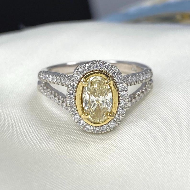 May include: A ring featuring a central oval-cut light yellow stone set in a yellow gold bezel, surrounded by a halo of small white stones. The band is made of silver-toned metal and features a split shank design encrusted with small white stones.