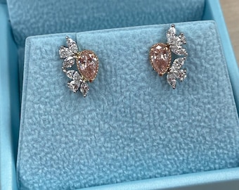 May include: A pair of elegant earrings featuring a pear-shaped, light pink gemstone surrounded by smaller, clear gemstones. The earrings are set in a silver metal and displayed on a light blue velvet surface inside a jewelry box.