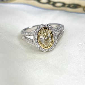 May include: A ring featuring a central oval-cut yellow stone set in a yellow gold bezel, surrounded by a halo of small white stones. The split shank band is crafted from silver-toned metal and is encrusted with rows of small white stones.