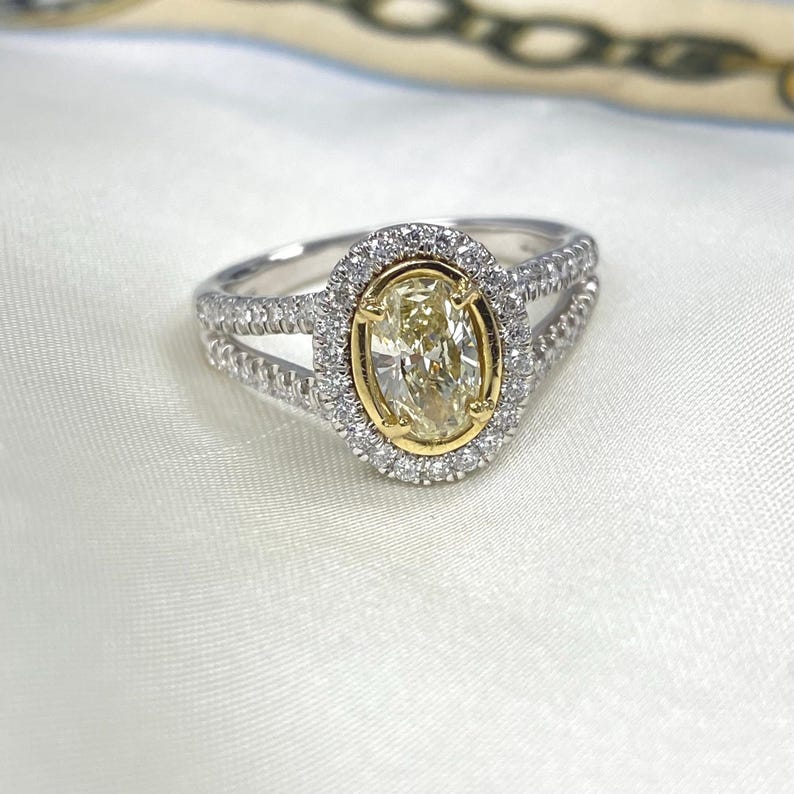 May include: A ring featuring a central oval-cut yellow stone set in a yellow gold bezel, surrounded by a halo of small white stones. The split shank band is crafted from silver-toned metal and is encrusted with rows of small white stones.