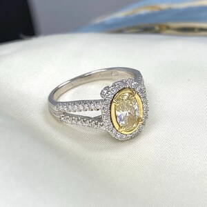 May include: A silver-toned ring featuring a central oval-cut yellow stone set in a yellow gold-toned bezel. The oval center is surrounded by a halo of small clear stones, with additional clear stones embedded along the split shank band.