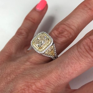 May include: A gold engagement ring with a large, square-shaped center diamond surrounded by a halo of smaller diamonds. The halo is accented with yellow diamonds. The ring is set in a white gold band.