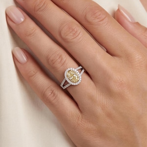 May include: A ring featuring a central oval-cut yellow stone surrounded by a halo of small white stones. The band is made of a silver-toned metal and splits into two rows near the setting, which are also encrusted with small white stones.
