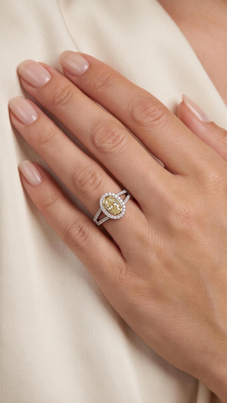 May include: A ring featuring a central oval-cut yellow stone surrounded by a halo of small white stones. The band is made of a silver-toned metal and splits into two rows near the setting, which are also encrusted with small white stones.