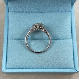 May include: A silver-toned ring featuring a central yellow gemstone surrounded by a halo of small white stones. The band is split and adorned with additional small white stones along the sides, presented in a light blue velvet box.