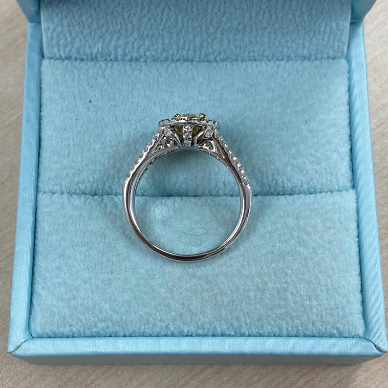 May include: A silver-toned ring featuring a central yellow gemstone surrounded by a halo of small white stones. The band is split and adorned with additional small white stones along the sides, presented in a light blue velvet box.