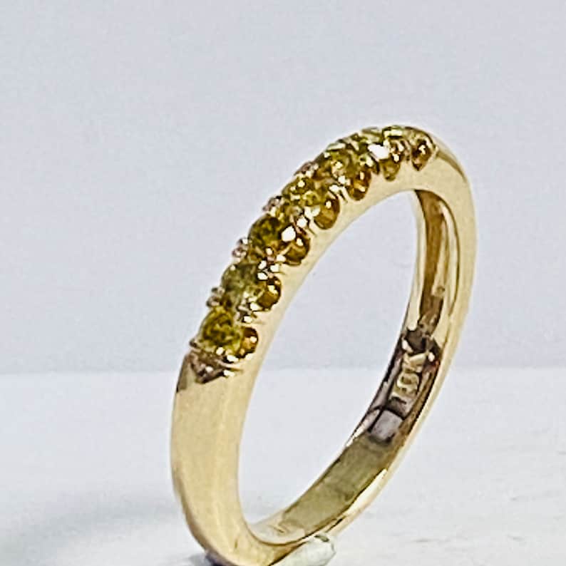 Puede incluir: Un anillo de banda de oro con una hilera de cinco peque&ntilde;as piedras preciosas redondas de color amarillo verdoso engastadas a lo largo de la curva superior. El interior del anillo tiene grabado 18K, indicando la pureza de esta joya de oro.