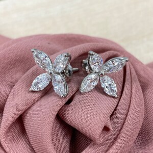 May include: A pair of silver earrings with four marquise-shaped diamonds set in a flower design. The earrings are on a pink fabric background.
