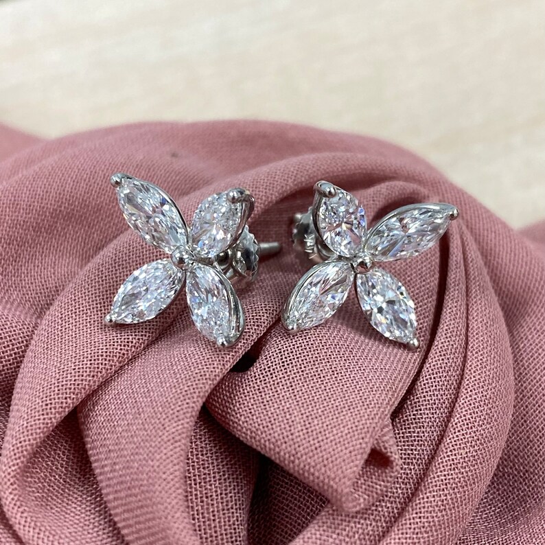 May include: A pair of silver earrings with four marquise-shaped diamonds set in a flower design. The earrings are on a pink fabric background.